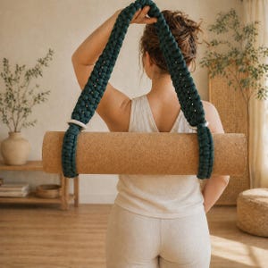 May include: A dark green macramé yoga mat carrier with white rings holds a rolled-up cork yoga mat. The carrier is held over the shoulder. The yoga mat is brown. The background includes plants and neutral tones.