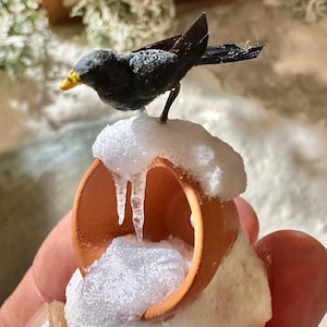 Op de afbeelding: Een miniatuur zwarte vogel met een gele snavel zit op een kleine, terracotta-kleurige pot bedekt met kunstsneeuw en ijspegels. De pot is gevuld met meer sneeuw, waardoor een winterse scène ontstaat.