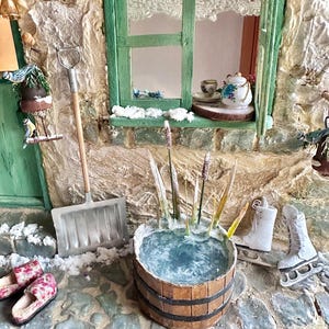 Könnte beinhalten: Miniatur-Winterszene mit einem rustikalen Steinhäuschen, einem grünen Fenster mit Spitzengardinen und einer Teekanne. Ein Holzfass mit Schilf und Eis steht neben Schlittschuhen und einer Schneeschaufel. Rosa Blumenslipper sind ebenfalls vorhanden.