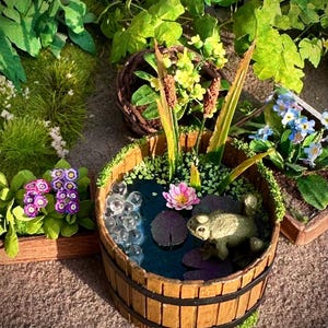 Könnte beinhalten: Ein Miniatur-Holzfass-Teich mit einer Froschfigur, Seerosen und Blasen. Der Teich ist von Grün und Blumen umgeben und schafft eine skurrile Gartenszene. Das Fass hat einen Durchmesser von etwa 15 cm.