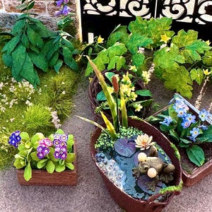 Könnte beinhalten: Detaillierte Miniaturgartenszene. Sie zeigt einen braunen bootsförmigen Teich mit einem Frosch, Seerosen und Wasserpflanzen. Weitere Elemente sind ein Holzpflanzgefäß mit lila Blumen und eine Backsteinmauer mit lila Blumen und grünem Laub.