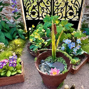 Könnte beinhalten: Miniaturgarten-Szene mit einem braunen Blumentopf mit Wasserspiel, einer rosa Lotusblume und einer Froschfigur. Weitere Miniaturpflanzen und -blumen befinden sich in Holzkisten und -körben. Im Hintergrund befindet sich ein schwarzes schmiedeeisernes Tor.