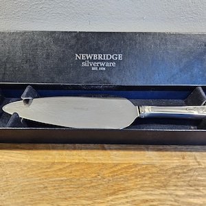 May include: A silver cake server with an ornate handle, presented in a black box with the "Newbridge Silverware" logo. The server has a wide, flat blade and is nestled in a blue velvet lining.