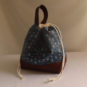 May include: A drawstring bag with a dark teal patterned fabric upper and a brown leather-look base. The bag features a brown handle and white drawstring closure. The pattern includes small white and orange shapes.