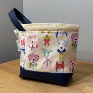 May include: A small, square tote bag with a navy blue base and handle. The main body of the bag is cream-colored and covered in a colorful pattern of various dog breeds. The top edge is a natural cream color.