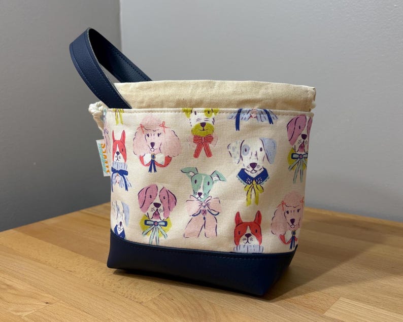 May include: A small, square tote bag with a navy blue base and handle. The main body of the bag is cream-colored and covered in a colorful pattern of various dog breeds. The top edge is a natural cream color.