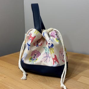 May include: A drawstring bag with a navy blue leather handle and base. The bag's cream-colored fabric is patterned with colorful cartoon dog faces. The drawstring is white, and the bag is sitting on a wooden surface.