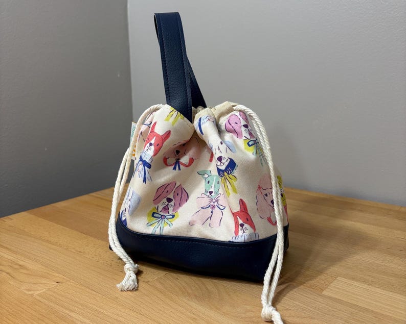 May include: A drawstring bag with a navy blue leather handle and base. The bag's cream-colored fabric is patterned with colorful cartoon dog faces. The drawstring is white, and the bag is sitting on a wooden surface.