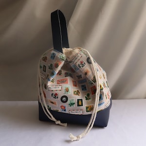 May include: A drawstring bag with a navy blue base and handle. The upper section features a cream background patterned with colourful postage stamp designs. It is tied with a white drawstring.
