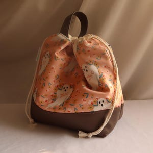 Peut inclure: Sac à cordon avec partie supérieure en tissu pêche ornée de fantômes blancs et de petites fleurs jaunes. Le bas est marron, avec une poignée marron et un cordon blanc. Le mot "boo" est visible sur les fantômes.