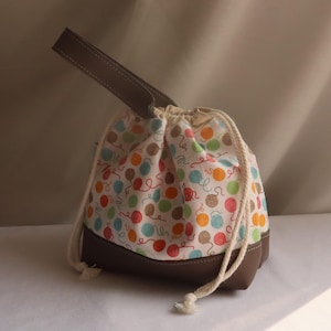 May include: A drawstring bag featuring a pattern of colourful yarn balls on a white background. It has a brown leather base and handle. The white drawstring bag is designed for carrying craft supplies, with a height of approximately 20 cm.