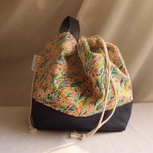 Peut inclure: Un sac à cordon floral avec une base bleu marine. Le sac a un cordon de serrage crème et une poignée bleu marine. Le motif floral comprend des fleurs rouges, jaunes et vertes. L'étiquette indique "MAKES".