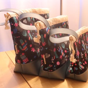 May include: Set of three black canvas tote bags with colorful cactus and geometric patterns. The bags have blue leather handles and a drawstring closure.