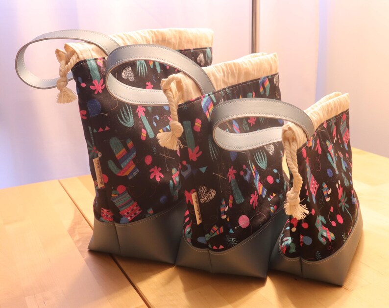 May include: Set of three black canvas tote bags with colorful cactus and geometric patterns. The bags have blue leather handles and a drawstring closure.