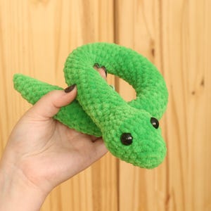 May include: A green crocheted snake toy with black eyes. The snake is curled up in a circle.
