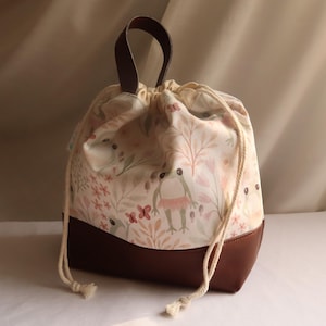 May include: A drawstring bag with a whimsical frog and floral print. The bag features a brown leather base and handle, with a cream-coloured drawstring closure. The bag's fabric has a light pink and green colour palette.