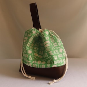 May include: A drawstring bag with a green and white pattern featuring gardening tools and vegetables. The bag has a brown leather bottom and handle, with a white drawstring closure.
