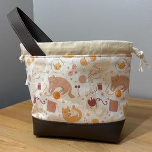 May include: A fabric project bag with a brown leather handle and base. The bag features a cream background with a repeating pattern of orange and brown cats, yarn balls, and sewing notions. It has a drawstring closure.