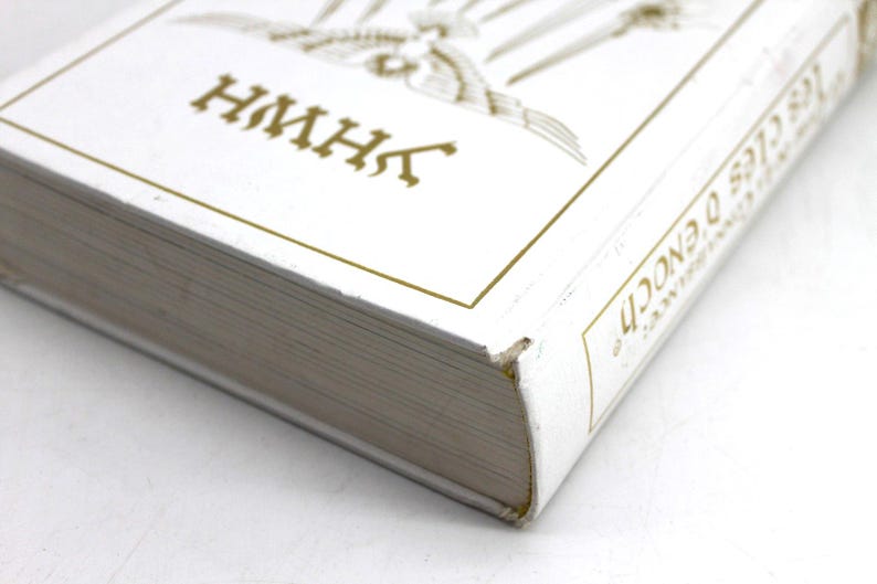 May include: A white book with gold lettering and accents. The title includes the text "Les Cl&eacute;s D'Enoch" and "HMHK". The book's pages are visible, and the cover has a gold border.