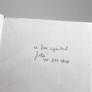 May include: Open book with a white textured page. Handwritten text in black ink reads "ce livre appartient Julie" followed by a phone number. The book's binding is visible on the left side.