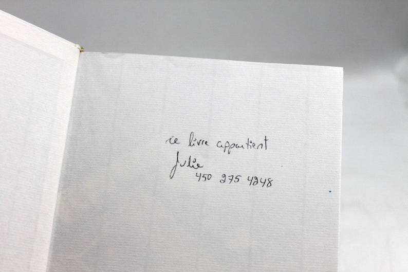 May include: Open book with a white textured page. Handwritten text in black ink reads "ce livre appartient Julie" followed by a phone number. The book's binding is visible on the left side.