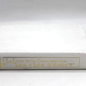 May include: A white book with gold lettering. The title "Le Livre de la Connaissance: Les Cl&eacute;s d'Enoch" is printed on the spine. The book is rectangular and appears to be in good condition.