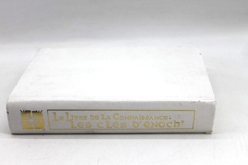 May include: A white book with gold lettering. The title "Le Livre de la Connaissance: Les Cl&eacute;s d'Enoch" is printed on the spine. The book is rectangular and appears to be in good condition.
