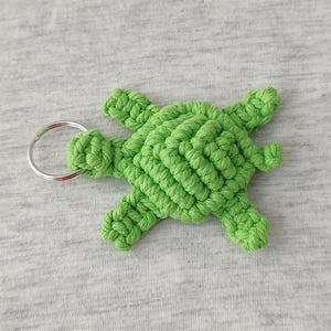 May include: A bright green macrame turtle keychain. The turtle is made of woven green cord and features a silver metal keyring. The turtle's body is a diamond pattern, and it has four flippers and a head.