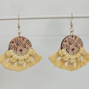 May include: A pair of handmade earrings featuring a light brown, semi-circular wooden pendant with a floral design. Below the pendant is a yellow macrame fringe. The earrings hang from gold-colored hooks.