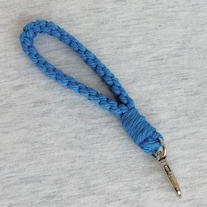 May include: A blue macrame wrist strap with a silver-colored clasp. The strap is made of braided cord and is looped to form a handle. The background is a light gray fabric.