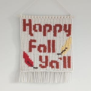 May include: A macrame wall hanging with the words "Happy Fall Y'all" in brown letters. The background is white with a red and yellow leaf design. The wall hanging is suspended from a wooden dowel with a white string.