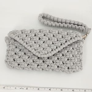May include: A light gray crochet clutch with a matching wrist strap. The clutch has a flap closure and a silver-colored clasp. The woven texture is visible throughout the bag. The bag is approximately 8 inches wide.