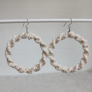 May include: A pair of cream-colored macrame hoop earrings. The earrings are made of twisted cotton rope, forming a circular shape. The earrings hang from silver-colored ear wires. The background is a light gray and white.