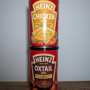 May include: Two cans of Heinz soup. The top can is red with white text and shows a bowl of chicken broth. The bottom can is red with white text and shows a bowl of oxtail soup. Both cans have the Heinz logo and the text "No artificial colours or preservatives."