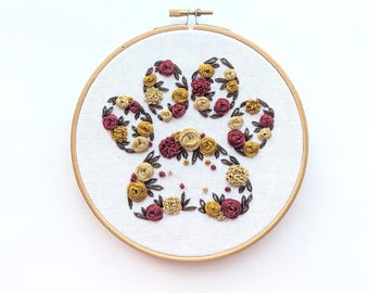 Könnte beinhalten: Ein floral besticktes Pfotenabdruck-Design in einem Holzreifen. Der Pfotenabdruck ist mit kleinen, detaillierten Blumen in Burgunder-, Gold- und Cremetönen gefüllt, vor einem weißen Stoffhintergrund. Der Holzreifen ist rund und hat einen Metallverschluss.