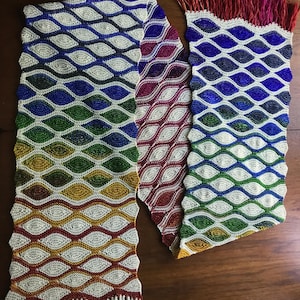 Puede incluir: Una larga bufanda de ganchillo de colores con flecos blancos en cada extremo. La bufanda está hecha con un patrón repetitivo de pequeños puntos en forma de lágrima. Los colores de la bufanda son rojo, morado, azul, verde, amarillo y blanco.