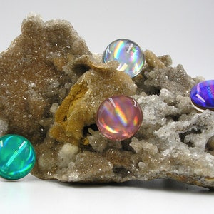 May include: Four iridescent glass cabochons in shades of blue, green, pink, and purple are displayed on a piece of rough, white, and brown rock.