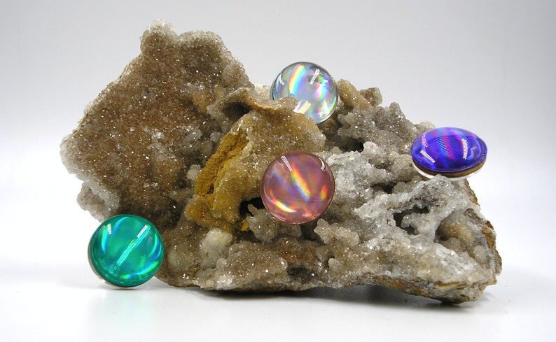 May include: Four iridescent glass cabochons in shades of blue, green, pink, and purple are displayed on a piece of rough, white, and brown rock.