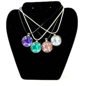 May include: Four round pendant necklaces with iridescent designs in shades of purple, green, and pink. The pendants are displayed on a black velvet jewelry display. Each pendant hangs from a black or white cord.