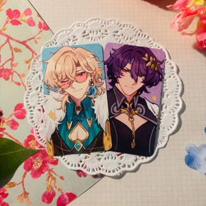 Puede incluir: Dos tarjetas de personajes ilustradas con hombres de estilo anime con cabello y ropa coloridos. El personaje de la izquierda tiene el cabello rubio, gafas de sol rosas y un atuendo azul y verde. El personaje de la derecha tiene el cabello morado, un atuendo negro y dorado y un collar de oro.