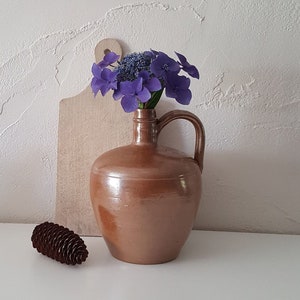 Può includere: Un boccale di ceramica marrone con manico contiene fiori viola. Sono visibili anche un tagliere di legno e una pigna marrone. Gli oggetti sono disposti su una superficie bianca contro un muro bianco strutturato.