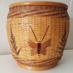 Puede incluir: Una cesta cilíndrica tejida con un diseño de mariposa y helecho. La cesta está hecha de material tejido marrón claro y marrón oscuro. La mariposa tiene un cuerpo rojo oscuro y alas marrones. Los helechos están en los lados.