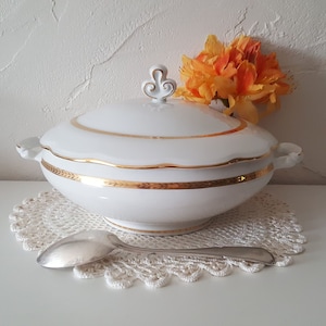 Peut inclure: Une soupière en porcelaine blanche avec un couvercle, ornée d'une bordure dorée et d'une poignée décorative. La soupière est posée sur un napperon en dentelle blanche, avec une cuillère en argent à proximité. Un arrangement floral orange est en arrière-plan.