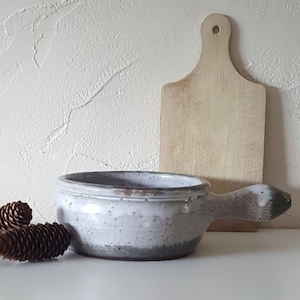 Op de afbeelding: Een gespikkelde witte keramische soepkom met een handvat, naast een houten snijplank en twee dennenappels. De kom heeft een grijze rand en bodem. De snijplank is lichtbruin.
