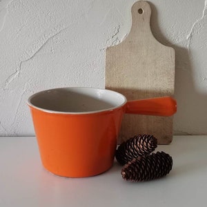 Può includere: Una casseruola in smalto arancione con interno bianco e un'unica maniglia. Un tagliere in legno e due pigne sono dietro la casseruola. Gli oggetti sono disposti su una superficie bianca contro un muro bianco strutturato.