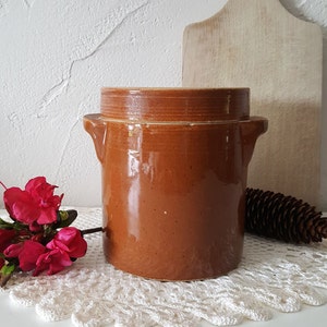 Peut inclure: Un pot en grès brun cylindrique avec un couvercle assorti et deux anses latérales. L'extérieur présente une finition lisse et brillante avec un léger mouchetage sur tout le corps en argile. Le récipient a un profil droit et un couvercle plat.
