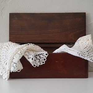 Peut inclure: Un coffret en bois brun foncé avec un couvercle à charnière, partiellement ouvert, révélant un napperon en dentelle crochetée blanche. Le napperon est drapé sur le bord du coffret, mettant en valeur son motif ajouré délicat.