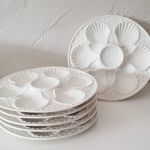 Könnte beinhalten: Eine Reihe weißer Keramik-Austernplatten mit Muscheldesign. Jede Platte hat sechs muschelförmige Fächer und eine zentrale Vertiefung. Die Teller haben einen strukturierten Rand und eine glänzende Oberfläche. Ein Stapel von Tellern ist sichtbar.