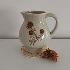 Peut inclure: Un pichet en céramique beige clair avec une finition mouchetée et une illustration botanique brune de fleurs de chardon sur le devant. Le pichet présente une anse incurvée et un bec verseur, posé sur un petit napperon en crochet crème.