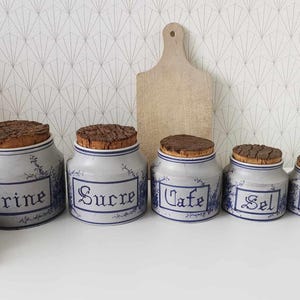 May include: A collection of five ceramic kitchen jars with cork lids. The jars are pale blue with blue lettering and floral patterns. The labels read "Farine", "Sucre", "Cafe", "Sel", and "Poivre". A wooden chopping board is behind them.